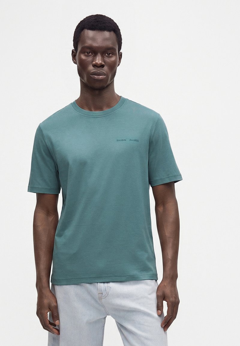 Homme portant un t-shirt à manches courtes bleu sarcelle et un jean bleu clair, debout devant un fond blanc uni.