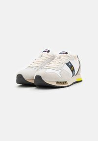 Sneaker con una toma in suede beige e mesh, suola in gomma nera, dettagli in blu navy e giallo, e un design testurizzato sulla suola.