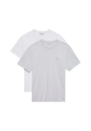 Jeune homme portant un sweat à capuche gris zippé, un t-shirt blanc, un pantalon large blanc et des sandales noires, debout les mains dans les poches sur un fond gris.