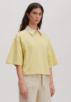 Woman with long dark hair wearing light yellow oversized short-sleeve shirt over white lace-trimmed pants, standing against plain background.