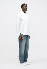 Fehér hosszú ujjú ing gombos gallérral és redős elemmel, világoskék bő szárú farmerrel és fehér tornacipővel párosítva.