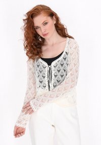 Wollene Strickjacke in Creme mit geometrischem Muster, offener Vorderseite und langen Ärmeln. Das Modell trägt darunter ein schwarzes Top und weiße Hosen.