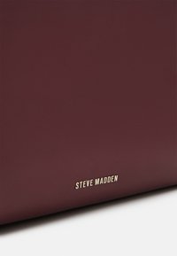 Sac à main en cuir bordeaux à texture lisse, avec un logo doré "Steve Madden" sur le devant. Design minimaliste, sans quincaillerie visible.