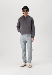 Grå hettegenser med frontlomme, lyseblå jeans og hvite joggesko med brune såler; modellen bruker svarte solbriller.