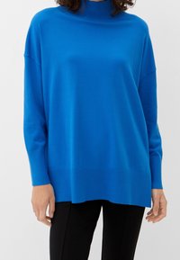 Personne portant un pull à col roulé ample de couleur bleu vif à manches longues et un pantalon noir, sur un fond blanc uni.