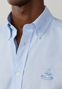 Gros plan sur le col d'une chemise à boutons bleu clair et le logo sur la poitrine avec une silhouette stylisée et le texte "FACONNABLE" sur le tissu.