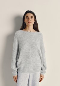 Woman with long dark hair wearing a loose light gray fuzzy sweater and light gray pants, standing against a plain beige wall.