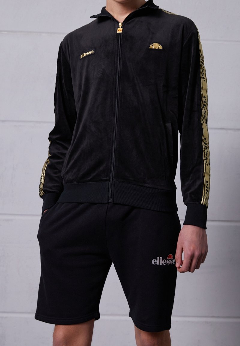 Svart velour joggjacka med guldaccenter och logotyp, tillsammans med svarta shorts med elastisk midja och sidofickor.