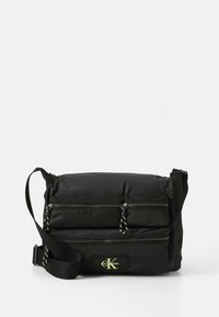 Mala tiracolo preta em nylon com dois bolsos frontais com fecho, alça ajustável e um patch de logótipo verde. Textura suave, formato retangular.