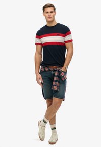 Navy and red striped short-sleeve t-shirt paired with denim shorts. Plaid shirt tied around the waist, white athletic shoes, and socks.