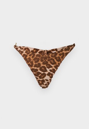 Kvinde iført leopardprint bikini og sorte højhælede sandaler, stående med hænderne løst foldet mod en ensfarvet hvid baggrund.