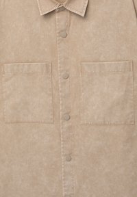 Beige színű, textúrált anyagból készült ing, amelynek gallérja, elöl gombos záródása van, és két egyenes szélű mellzsebe, amelyekhez illeszkedő gombok tartoznak.