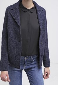 Giacca blu in misto lana con rever a punta, due bottoni frontali e tasche laterali, abbinata a una blusa nera e jeans blu.