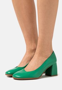 Talons hauts en cuir vert avec un nez arrondi et un talon épais, présentant une texture lisse et un design courbé.