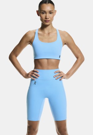 Frau steht mit den Händen in die Hüften gestützt, trägt ein hellblaues Sport-BH und hoch taillierte Radlerhose vor weißem Hintergrund.