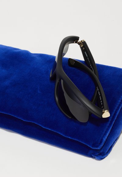 Ochelari de soare negri cu finisaj mat, așezați pe un sac cu catifea albastră texturată. Prezintă accente aurii pe cadru și lentile închise.