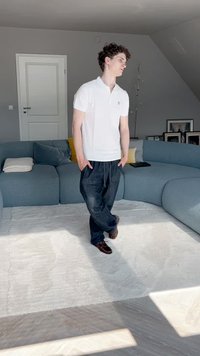 Biała koszulka polo, ciemne luźne spodnie i brązowe buty. Stoi na jasnym dywanie w nowoczesnym pomieszczeniu z zakrzywioną niebieską kanapą i neutralnymi ścianami.