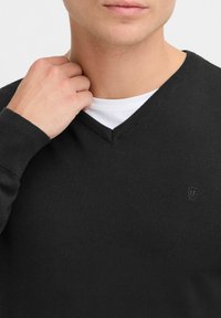 Black v-neck sweater made of knit fabric, featuring ribbed cuffs and a subtle logo on the chest. Worn over a white t-shirt.