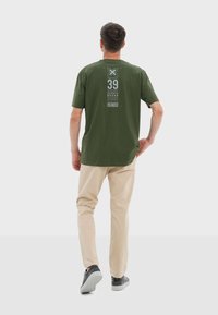Camiseta de manga corta verde oliva con estampado gráfico blanco y texto en la parte de atrás, combinada con pantalones beige y zapatillas grises.