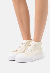 Beige högkoniga sneakers tillverkade av texturerad tyg med vita gummisulor och snörning. Har en upphöjd häl och subtila designaccenter.