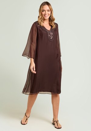 Mujer sonriente con un vestido marrón oscuro transparente hasta la rodilla con bordados en el escote en V y mangas abiertas tres cuartos, combinado con sandalias marrones.