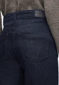 Temno modre kavbojke iz jeansa imajo visok pas in dva zadnja žepa. Tkanina je gladke teksture, dopolnjena z usnjenim znamčnim obližem.