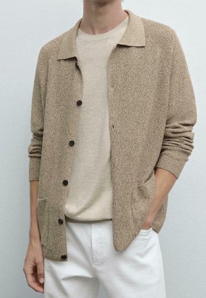 Mann trägt beige texturierten Knopfstrickcardigan über einem hellbeigen Hemd und weißen Hosen, mit einer Hand in der Tasche.