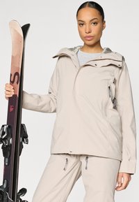 Geacă de schi impermeabilă bej, cu buzunar cu fermoar pe piept, glugă atașată și manșete reglabile, asortată cu pantaloni de schi.
