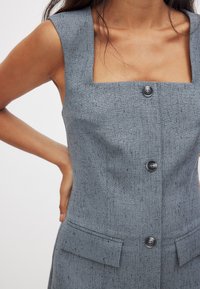 Pilkas tekstūruotas suknelė su kvadrato formos iškirpte, trumpomis rankovėmis, dviem priekinių kišenėmis ir juodais mygtukais viduryje.