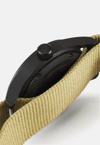 Crni sat s teksturiranim okruglim kućištem, izraženom krunicom i bež najlonskim remenom, na kojem je prikazan krupni plan bočnog profila sata.