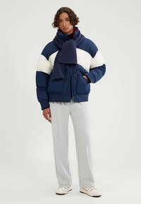 Marineblaue und weiße gesteppte Jacke mit hohem Kragen, großen Fronttaschen und passendem Schal. Hellgraue Hosen und cremefarbene Schuhe vervollständigen das Outfit.
