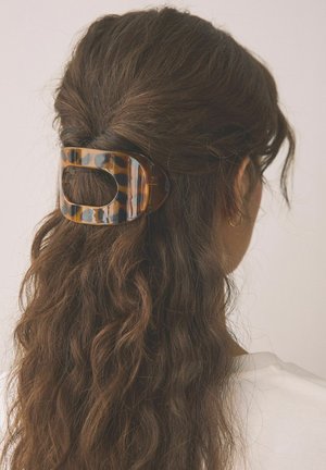 Braune, wellige Haare, halb zurückgesteckt mit einer großen rechteckigen Schildpatt-Haarklammer, Person trägt goldenen Creolen-Ohrring und weißes Oberteil.