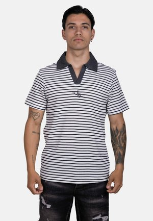 Joven con una camisa polo blanca y negra a rayas con cuello oscuro, de pie con las manos relajadas, mostrando tatuajes en ambos brazos.