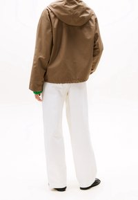 Bruine capuchonjas met een losse pasvorm, gecombineerd met witte wijd uitlopende broek en zwarte schoenen, gezien van achteren. Soepele textuur, casual ontwerp.