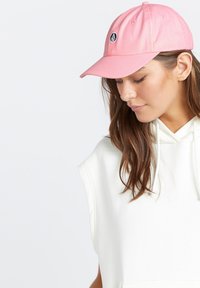 Roze pet met een ronde klep en geborduurd logo aan de voorkant. Draag het met een witte mouwloze hoodie voor een ontspannen uitstraling.