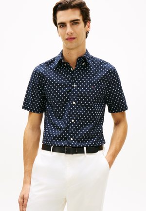 Mladý muž nosiaci tmavomodrú krátku košeľu na zapínanie s bielym vzorom, ktorá je zasunutá do bielych nohavíc s čiernym opaskom.
