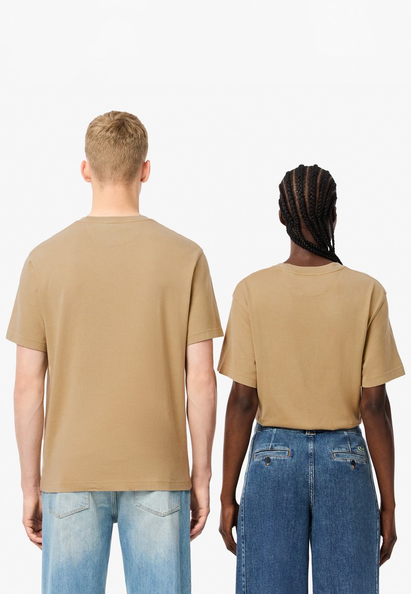 Zwei Personen, die identische hellbraune, kurzärmelige T-Shirts tragen und Rücken an Rücken stehen; beide T-Shirts sind schlicht, locker geschnitten und haben einen runden Halsausschnitt.