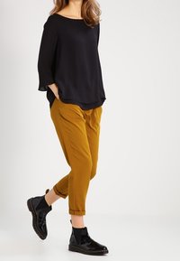 Haut en crêpe noir à manches larges, pantalon moutarde fuselé, et bottines noires brillantes à semelle épaisse avec des coutures décoratives.