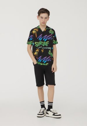 Maglietta nera a maniche corte con testo colorato "SURF" e disegni di palme, abbinata a pantaloni corti neri arrotolati e sneakers bianco e nere.