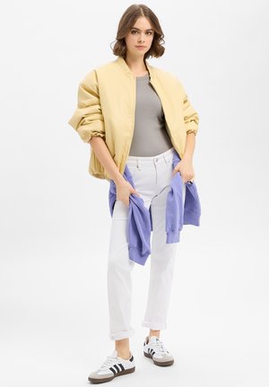 Jeune femme debout portant une veste bomber jaune, un haut gris, un jean blanc, des baskets blanches, et tenant un sweat-shirt violet plié.