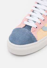 Gros plan d'une basket avec des lacets blancs, un embout bleu, des côtés roses et un détail jaune sur une semelle en caoutchouc blanche.