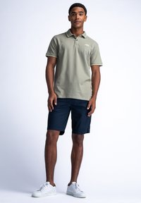 Kortärmad olivgrön polo med krage och subtila randiga detaljer, ihopparad med marinblå shorts och vita sneakers. Slät tyg, avslappnad passform.