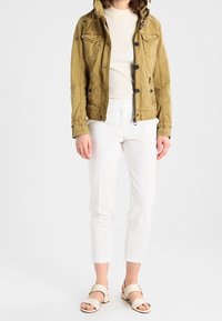 Olivgrön jacka med knäppbara fickor, krämfärgad ribbad topp, vita avsmalnande byxor och beige sandaler med spännen. Avslappnad utomhusoutfit.