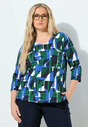 Kvinde med langt blondt hår iført olivengrønne briller og en top med geometrisk mønster i blå, grøn, sort og hvid samt sorte bukser.