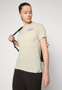 Light beige short-sleeve t-shirt with purple graphic text on the chest, paired with dark pants and a black backpack. Soft, textured fabric.