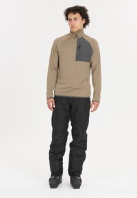 Brauner Langarm-Pullover mit einer dunkelgrauen Brusttasche; kombiniert mit lockeren schwarzen Hosen und schwarzen Schneestiefeln. Durchgehend glatte Textur.