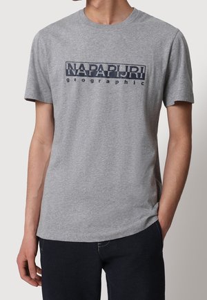 Mężczyzna noszący jasnoszary t-shirt z krótkim rękawem z logo „NAPAPIJRI geographic” w ciemnoniebieskim kolorze, w połączeniu z ciemnymi spodniami.