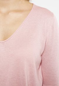 Vaaleanpunainen, pehmeä neulepaita, jossa on syvä V-pääntie. Puserossa on hienovaraista ribbitekstuuria pääntiellä ja hihoissa, mikä korostaa sen rakennetta.
