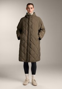 Olijfgroene, gewatteerde lange jas met een capuchon, voorzien van een rits aan de voorkant, zijzakken en een relaxte pasvorm. Draagt over zwarte legging en sneakers.