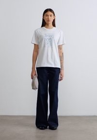 Λευκό βαμβακερό t-shirt με γραφική εκτύπωση, συνδυασμένο με σκούρο μπλε τζιν με φαρδιά γραμμή. Το μοντέλο κρατά μια μικρή ασημένια τσάντα, στέκεται σε ουδέτερο φόντο.